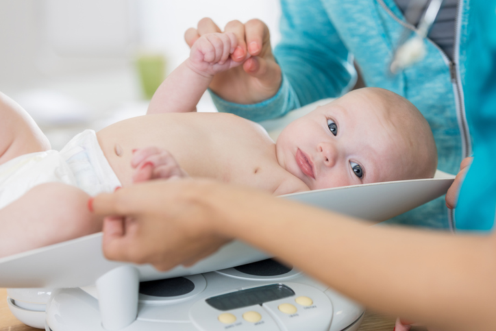 An unrecognizable pediatric nurse weighs a baby girl during well check appointment. The baby's mother holds the adorable girl's hand.