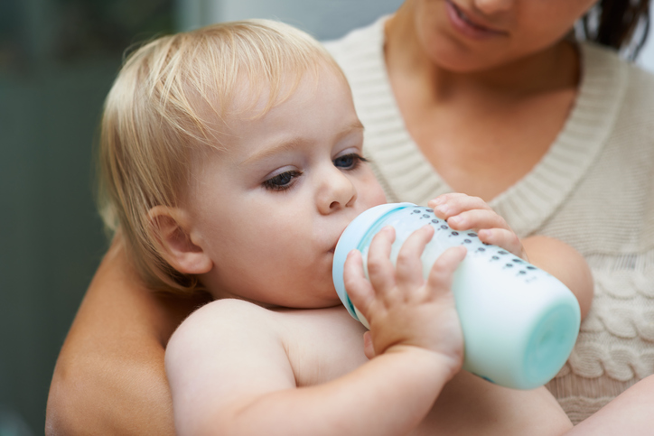 A baby boy drinking milk from his bottle while being held by his mother