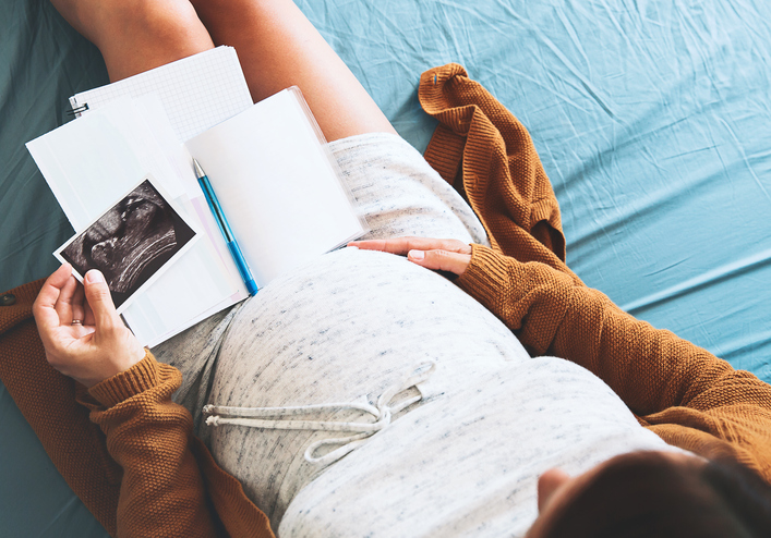 Pregnant woman makes notes, looking at ultrasound image and medical documents. Concept of pregnancy, health care, gynecology, medicine. Mother waiting of baby. Close-up, copy space, indoors.