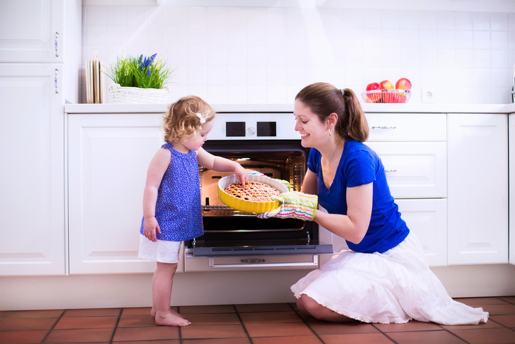 Mother and child bake a pie. Young woman and her daughter cook in a white kitchen. Kids baking pastry. Children helping to make dinner. Modern interior with oven and other appliances. Family eating.