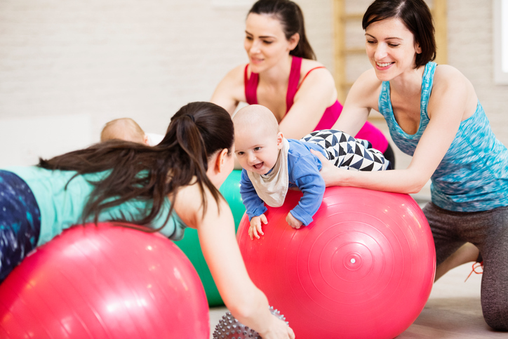 Group of fit young mothers and their babies in modern gym with personal trainer doing exercises on gymnastic balls. Healthy lifestyle.