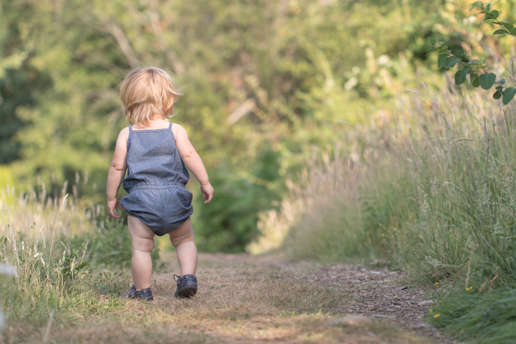 Blond baby in blue jeans costume walking away down on green path, baby's first steps