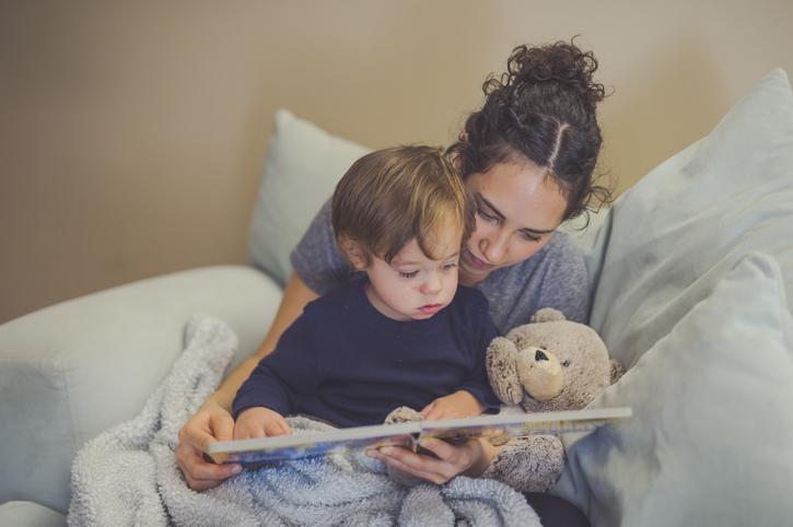 Young Hispanic mom reads a book aloud to her toddler son. They're pointing at the pictures together. A teddy bear is snuggled up with them.