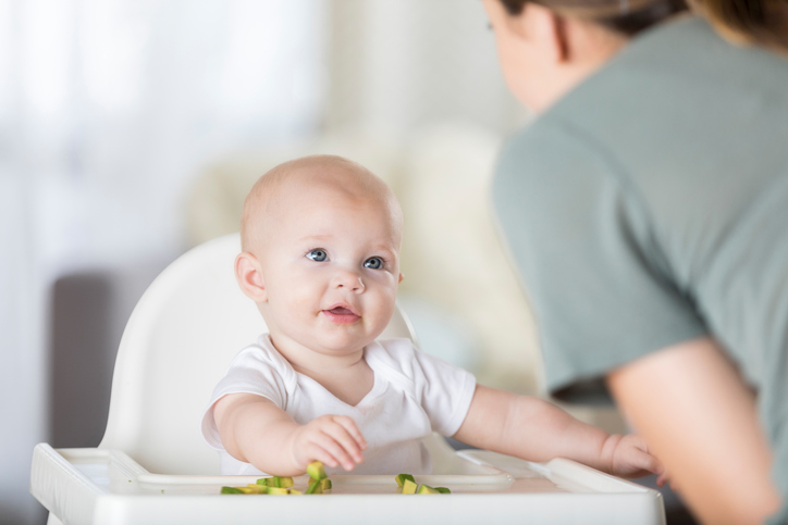 Beautiful baby girl looks at her mom. The baby is eating an avocado as she sits in her high chair at home.