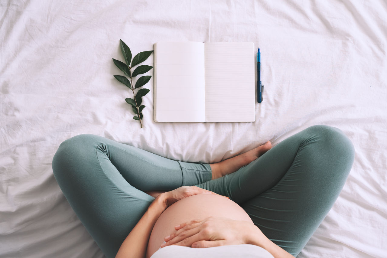 Pregnant woman with beautiful belly makes notes or check list in paper diary. Concepts of preparation for baby birth, tips for a healthy pregnancy. Minimalist style photography. Close-up, indoors.